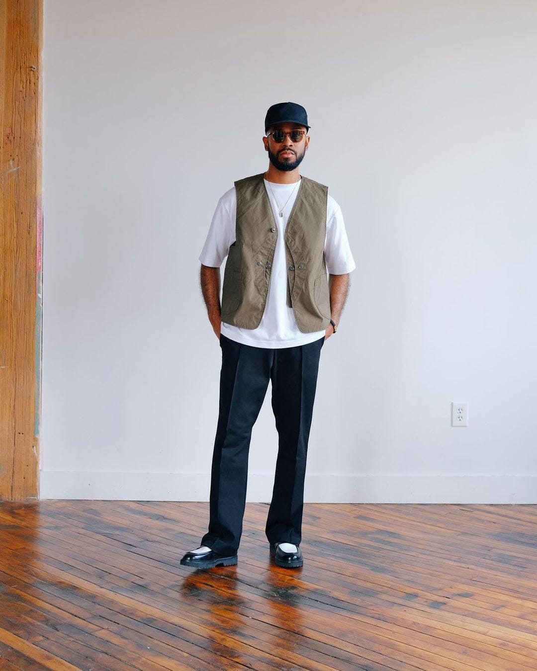 man standing on a wood floor in front of a white wall wearing a white t-shirt with tan v-neck vest and dark pants with black and white penny loafers