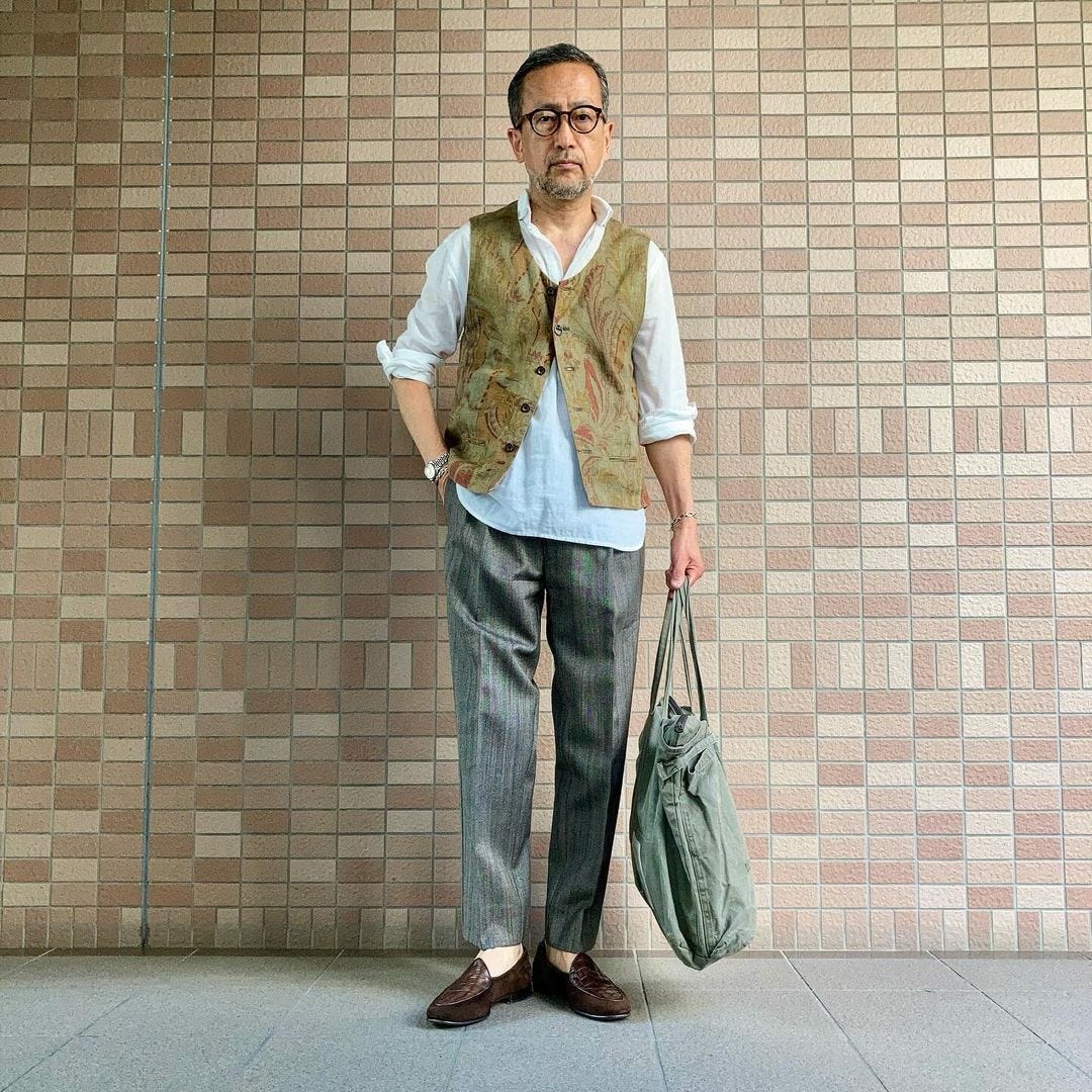 man standing in front of a brick wall wearing a brocade vest over a white collared shirt with grey trousers and brown loafers, carrying a large tote bag