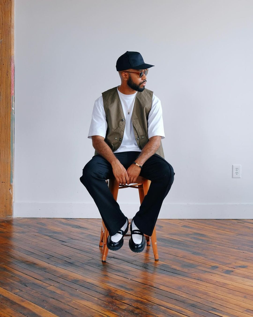 man sitting on a stool wearing a white t-shirt with tan v-neck vest and dark pants with black and white penny loafers