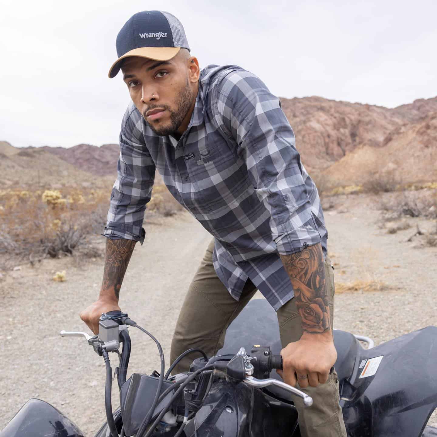 man standing on an ATV wearing a checked shirt and a cap