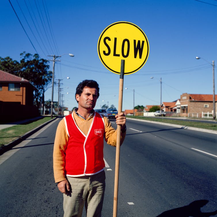 Man standing on a street, wearing orange 'hi-vis' vest holding a yellow sign that says 'slow'