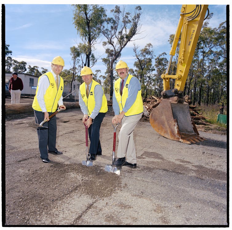 Three politicians wearing business wear and yellow hi-vis vests, with silver shovels, about to dig into dirt at a new construction site