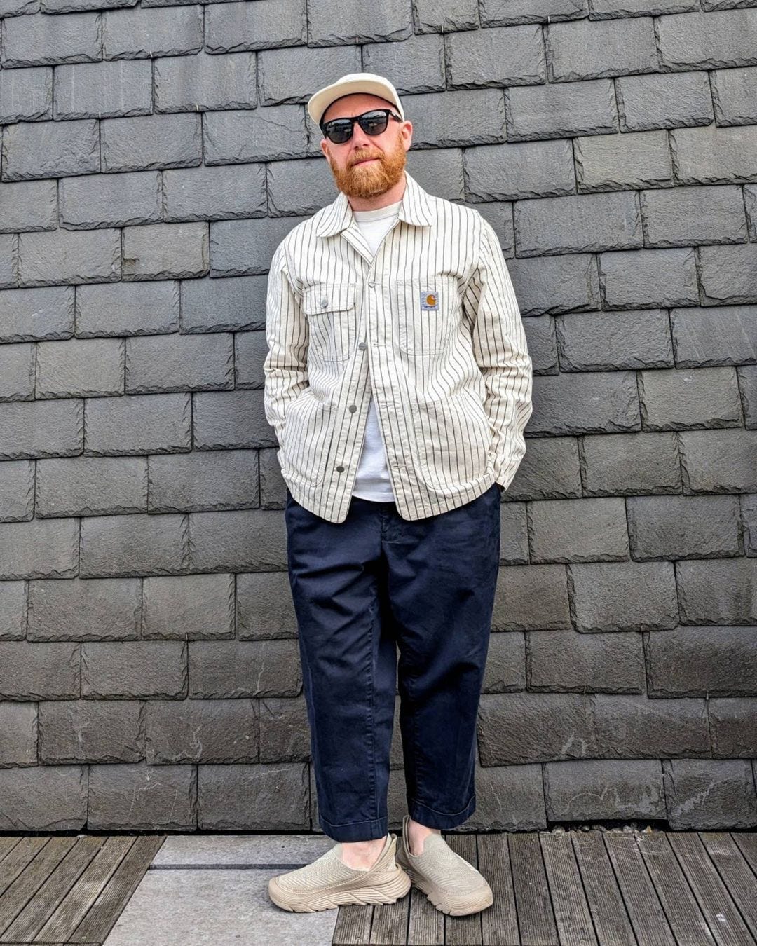 man standing in front of a brick wall wearing a pinstripe carhartt jacket with navy pants and a baseball hat