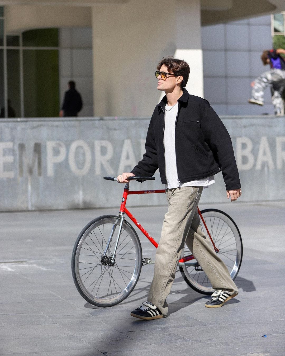 man walking with a red bicycle, wearing a black workwear jacket over a white shirt with tan pants and sneakers