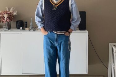 bearded man wearing a navy sweater vest over a light blue collared shirt with jeans and grey Converse sneakers