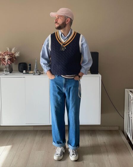 bearded man wearing a navy sweater vest over a light blue collared shirt with jeans and grey Converse sneakers