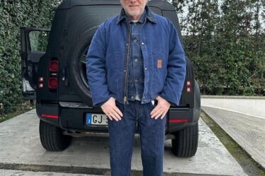 older man standing in front of a black car in a driveway, wearing a blue carhartt jacket over a blue shirt with blue jeans