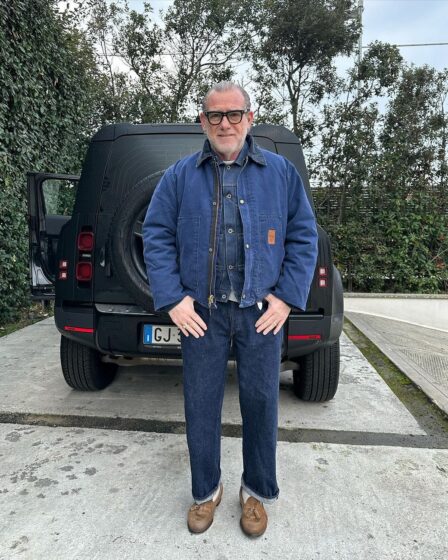 older man standing in front of a black car in a driveway, wearing a blue carhartt jacket over a blue shirt with blue jeans