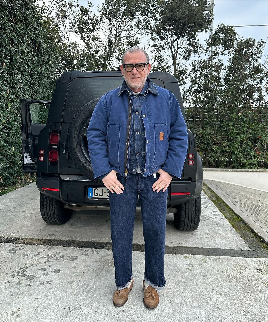 older man standing in front of a black car in a driveway, wearing a blue carhartt jacket over a blue shirt with blue jeans