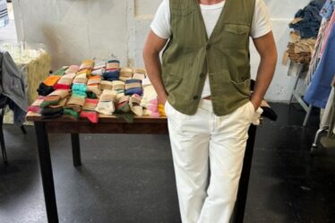 man leaning against a table full of socks, wearing an olive green vest with tan pants and suede birkenstock boston clogs with red socks