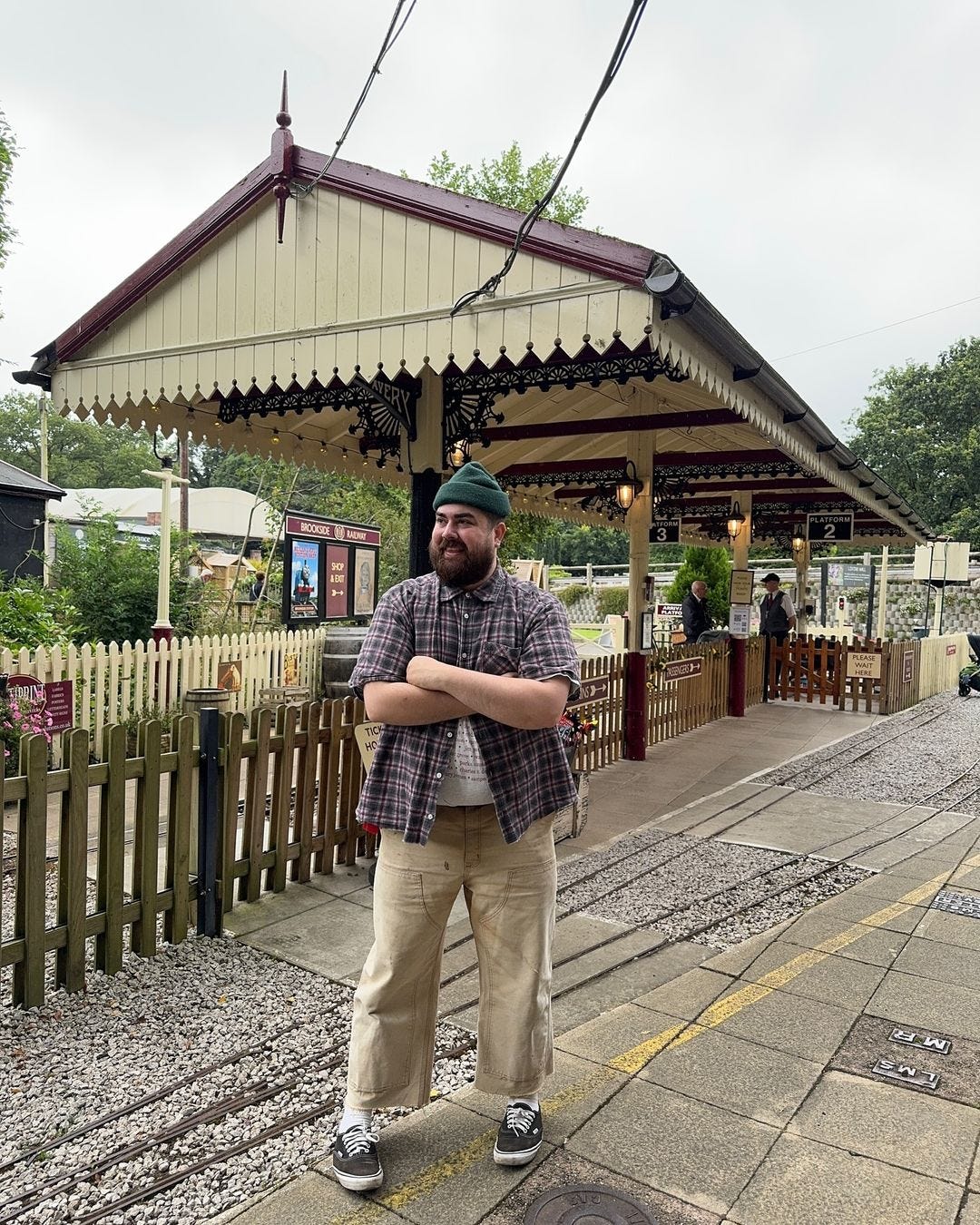 man standing at a train station wearing a short-sleeve plaid shirt with ankle-baring khaki-colored pants