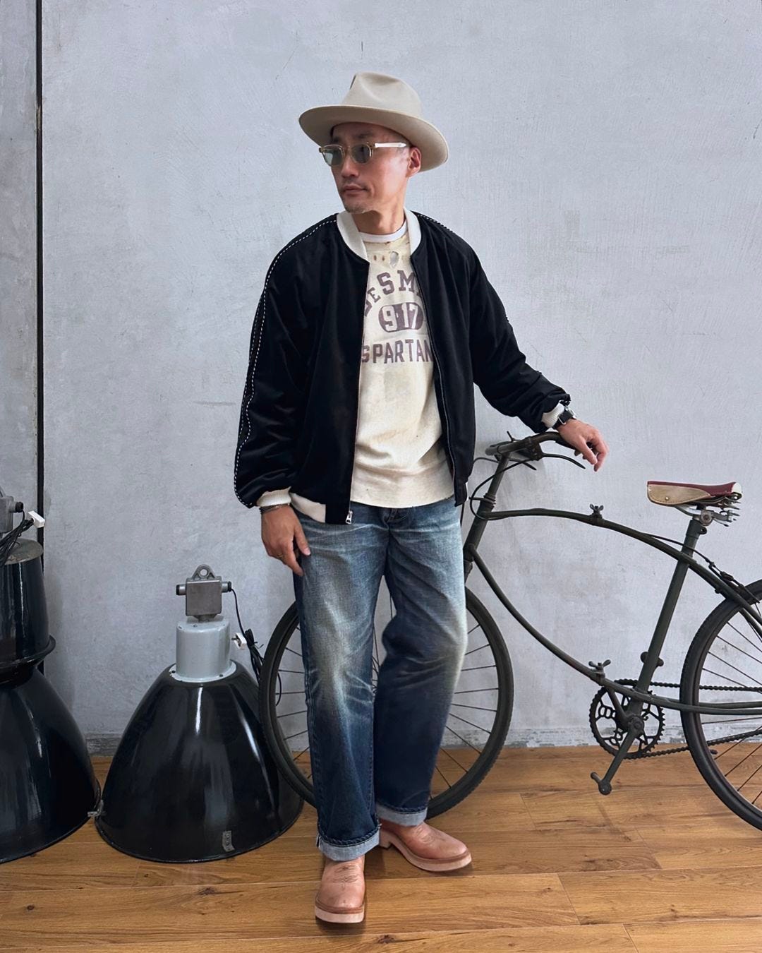 man standing inside next to a bike, wearing a navy bomber jacket, western-style hat, sweatshirt and jeans