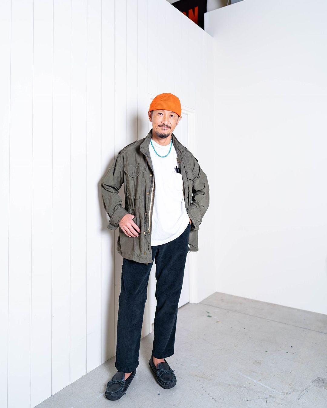 man standing in a corner wearing an orange hat, white shirt, olive jacket, turquoise necklace, dark pants and loafers, men's fall date outfits