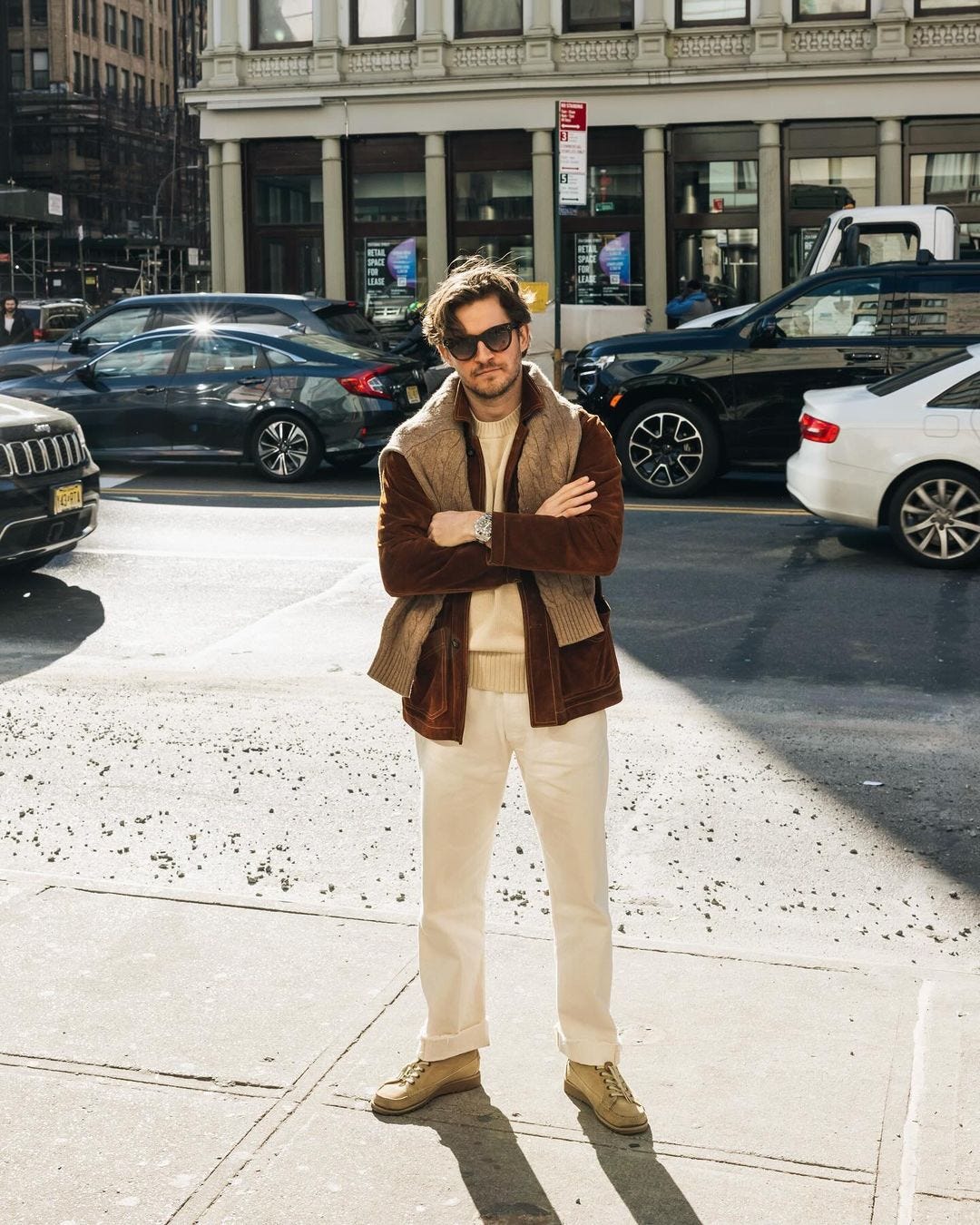 man standing on a sidewalk in sunglasses, wearing a brown coat with a tan sweater over the shoulders, light-colored pants, and suede shoes