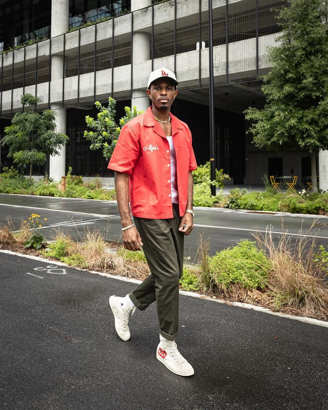 man walking on the street wearing a red short-sleeve shirt over a white tank with olive green pants and high-top nike sneakers