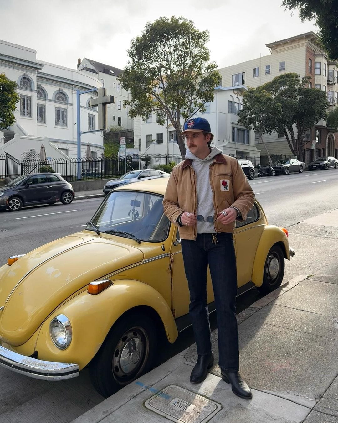 man standing in front of an old yellow car wearing a blue baseball cap, grey hoodie, tan workwear jacket with a rose embroidered on the chest pocket,dark jeans, and black cowboy boots