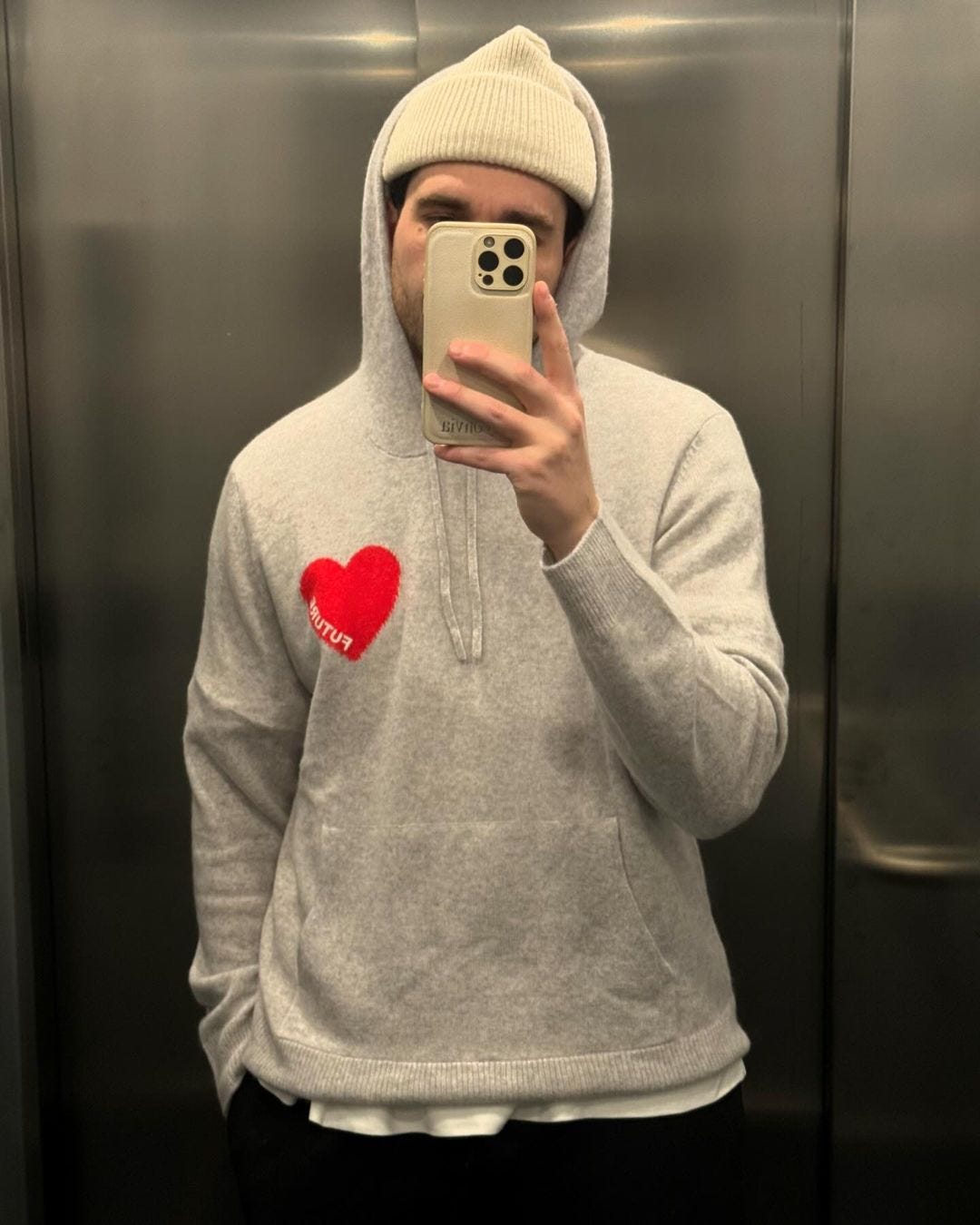 man taking a selfie wearing a grey knit hoodie with a red heart on the chest and a cream-colored beanie