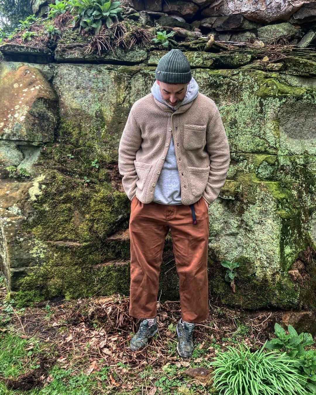 man standing outside in front of mossy stones wearing a grey beanie, tan sherpa jacket, grey hoodie sweatshirt, brown pants,and camo sneakers