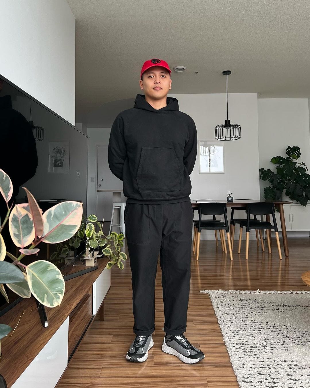 manstanding in a living room wearing all black with a red baseball cap