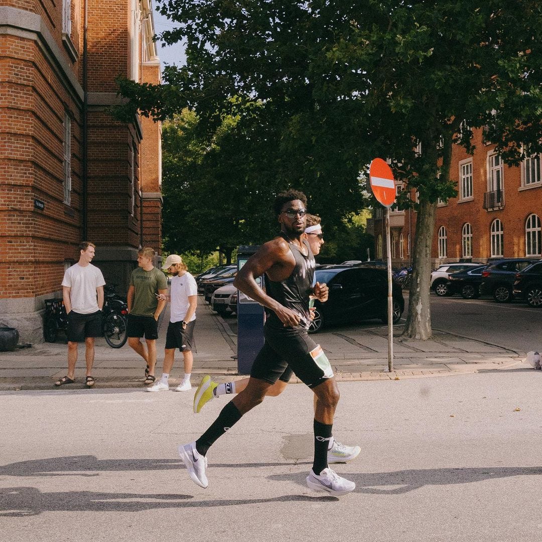 two athletic men running in a race crossing a street in front of three casually dressed young men