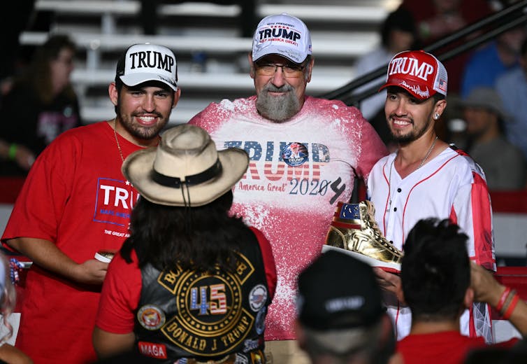 Three men wear white and red shirts that say 'Trump' and hats that also say 'Trump.' A person wearing a cowboy hat faces them and they smile. One of the men holds a pair of gold high top sneakers.