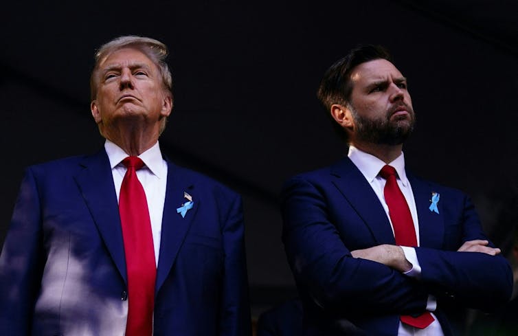 Two white men wear dark navy suits, red ties and white dress shirts and they have serious faces on.