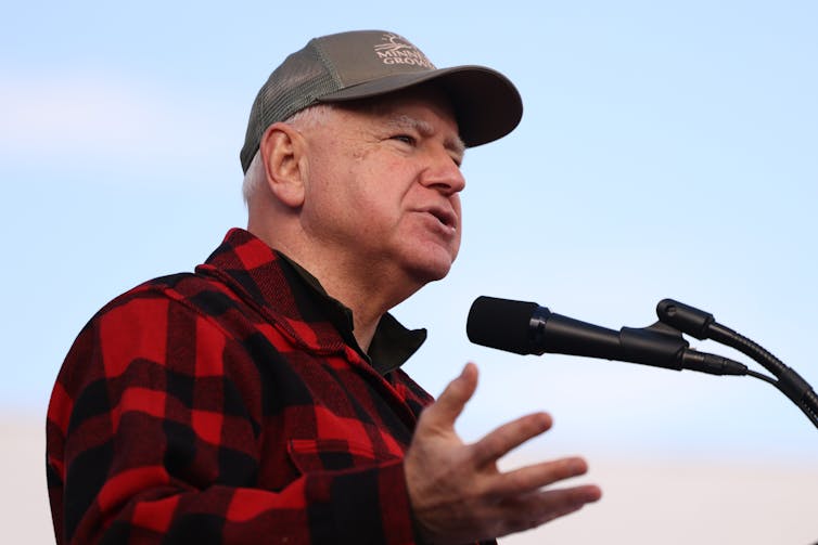 A white man wears a black and red flannel shirt and a hat and speaks into a microphone.