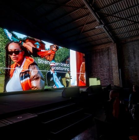 A dark room with an audience and a big screen with a fashion campaign image with "strengthening brand positioning" on the screen.