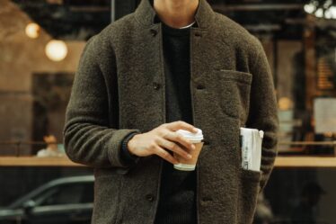 man wearing a boiled wool blazer over a crewneck sweater with a baseball hat, holding a cup of coffee