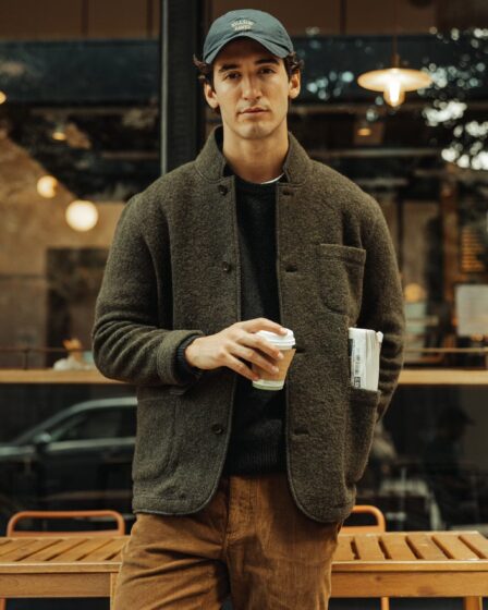 man wearing a boiled wool blazer over a crewneck sweater with a baseball hat, holding a cup of coffee