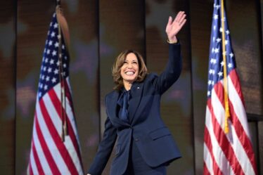 A woman wears a dark blue pantsuit and waves her hand and smiles. She stands in between two American flags.