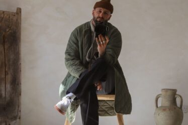 man sitting on a wooden stool in a minimally decorated room taking a selfie, wearing a brown beanie, green long quilted jacket, dark pants, and white sneakers | men's long coat outfits