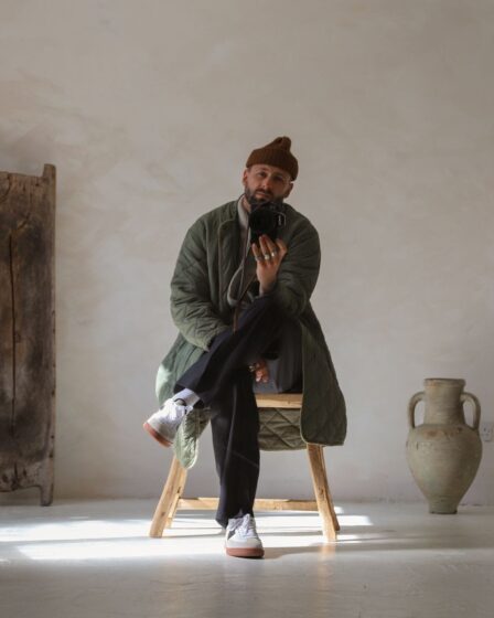 man sitting on a wooden stool in a minimally decorated room taking a selfie, wearing a brown beanie, green long quilted jacket, dark pants, and white sneakers | men's long coat outfits