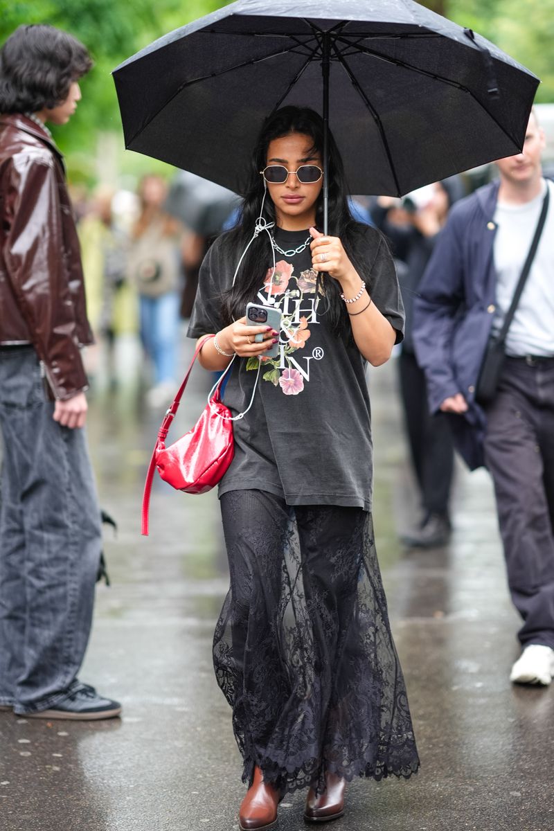 A guest wears silver rimmed with black lenses sunglasses, silver chain necklace, gray oversized floral/print shirt with white letters, shiny red leather bag, dark gray skin tight form fitting pants, black lace sheer mesh maxi skirt, shiny light brown boots leather shoes, white beaded bracelet, white bracelet, gold ring, during the Paris Fashion Week Menswear Spring/Summer 2025 on June 21, 2024 in Paris, France. (Photo by Edward Berthelot/Getty Images)