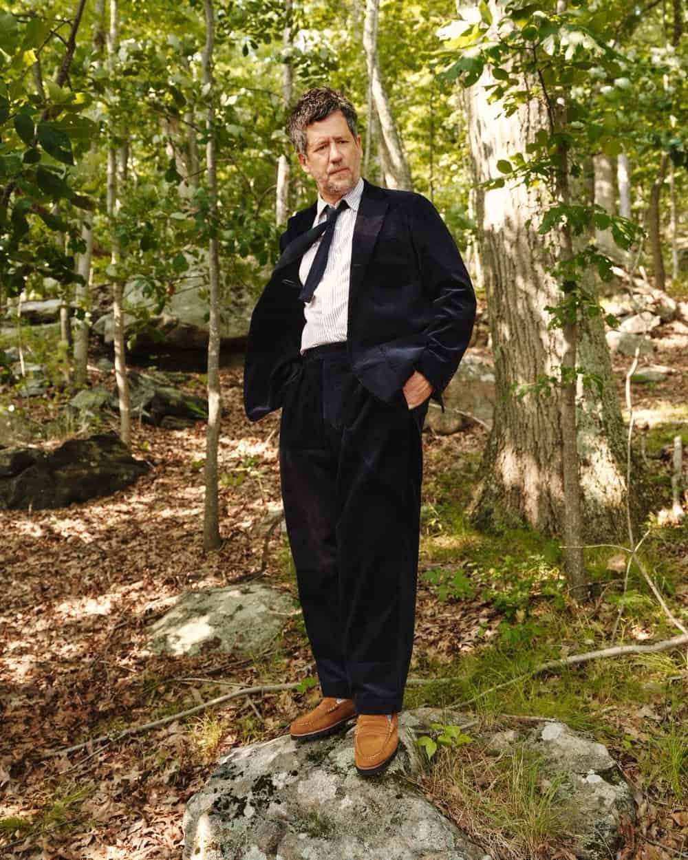 man wearing a black cord suit and brown loafers while standing on a large rock