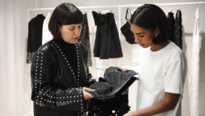 Two designers study a piece of material, with a rack of clothing displayed behind them.