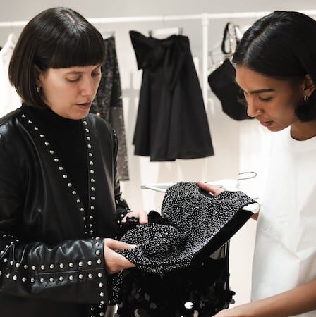 Two designers study a piece of material, with a rack of clothing displayed behind them.