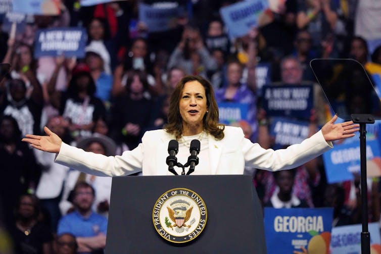 Vice-president Kamala Harris in a white suit speaking to a large crowd, arms outstretched.