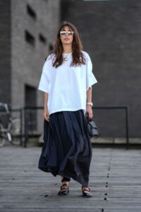 A guest wears light brown rimmed sunglasses, gold chain with gold heart shaped pendant, white oversized t-shirt, a silver watch, black shiny leather bag, black flowy maxi skirt, shiny black Mary Janeâs leather shoes, outside Forza Collective, during the Copenhagen Fashion Week Spring/Summer 2024-2025 on August 5, 2024 in Copenhagen, Denmark. (Photo by Edward Berthelot/Getty Images)