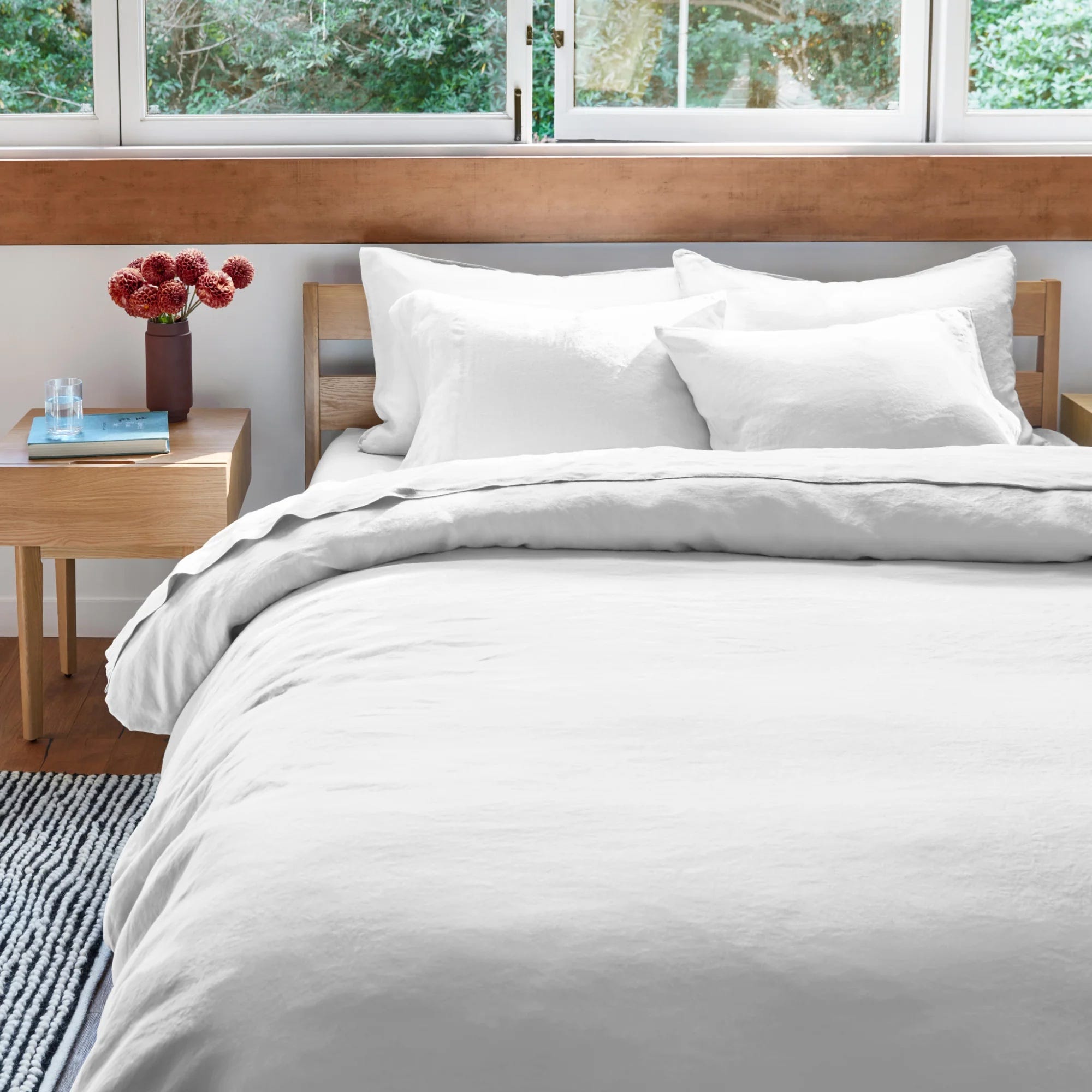 well-designed bedroom with bed made with white linen bedding next to a wood nightstand in front of wall with windows looking out to trees outside