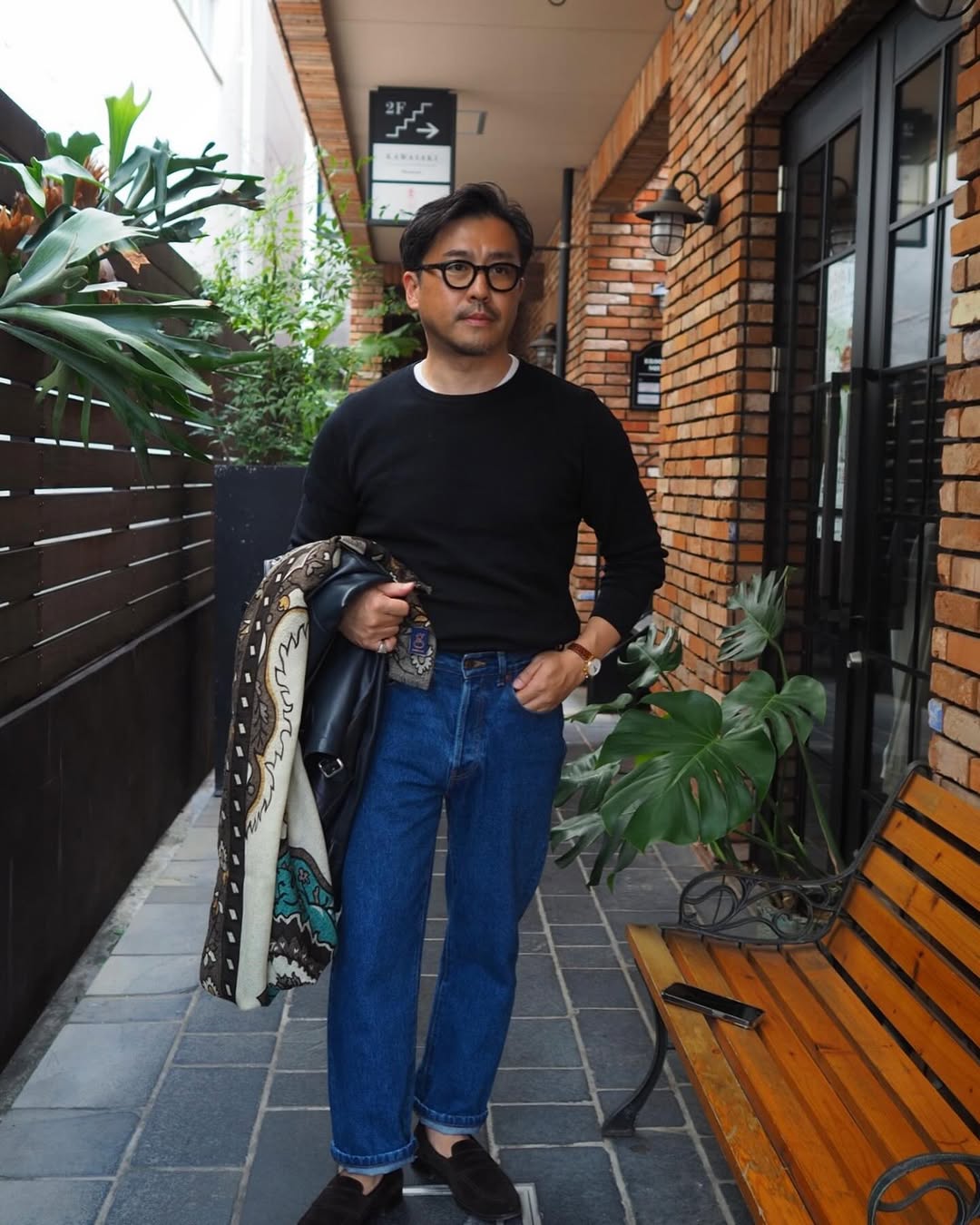 stylish man wearing blue jeans and a black sweater, holding a black leather jacket and silk patterned scarf in one arm