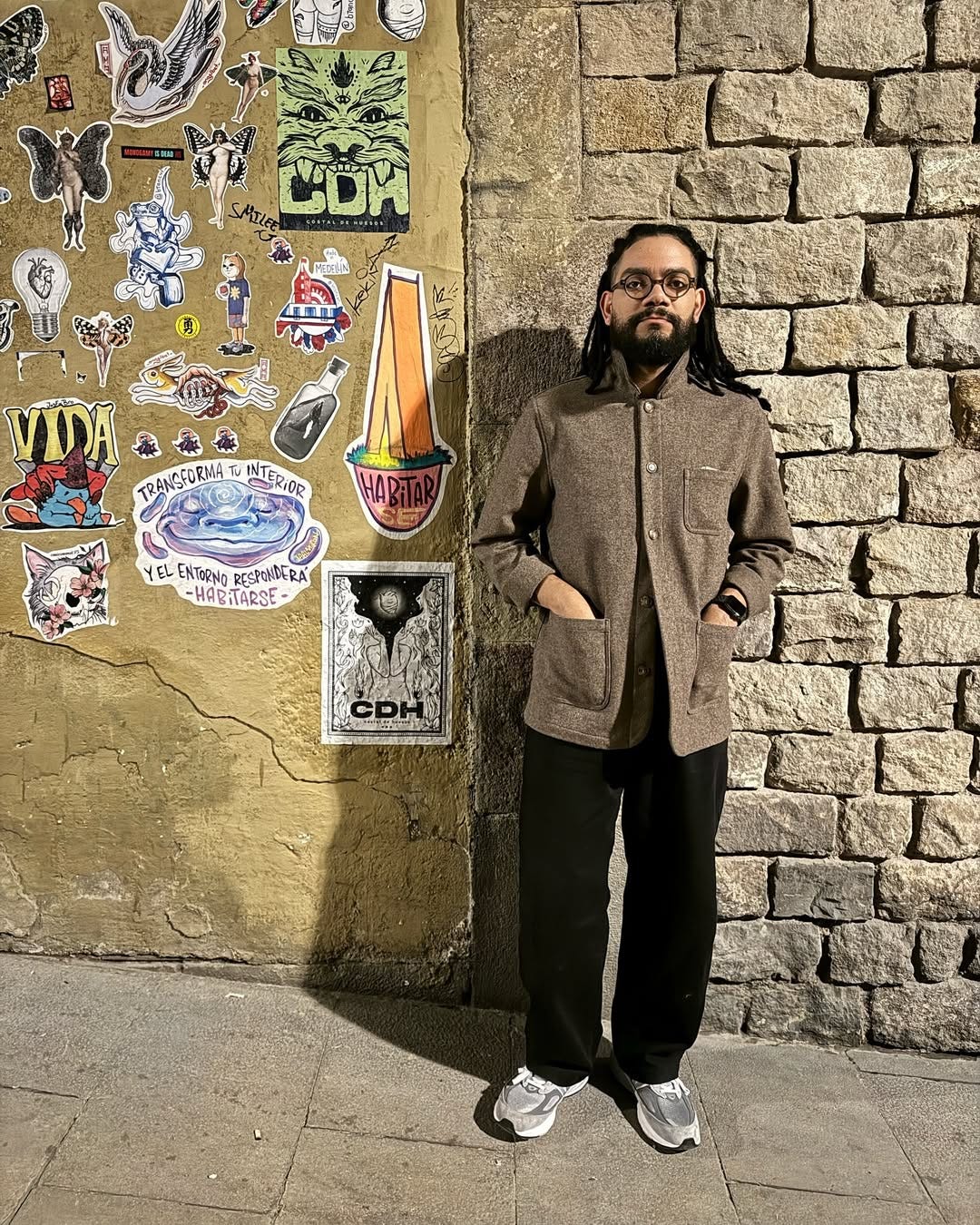 stylish man standing outside in front of a wall with stickers on it wearing a brown chore coat buttoned at the top with wide leg black pants and grey New Balance sneakers