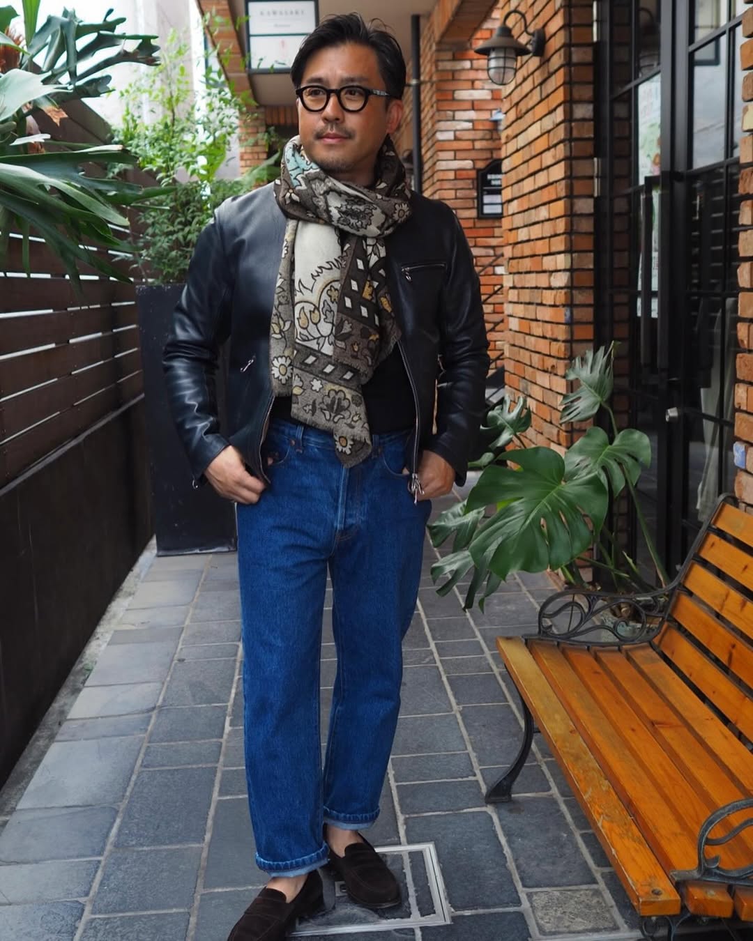 stylish man wearing blue jeans, a black sweater, black leather jacket, and silk patterned scarf