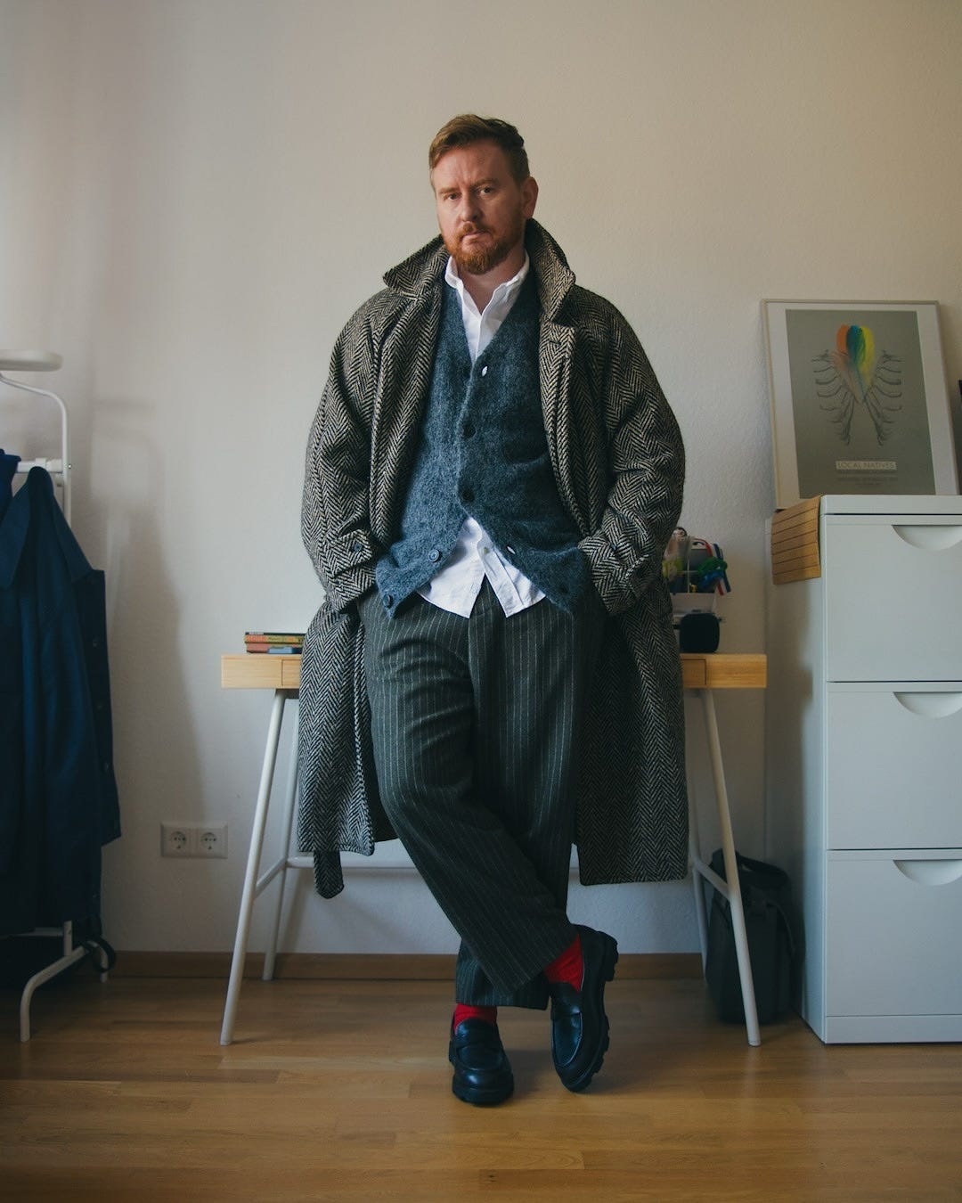 stylish man wearing a long herringbone topcoat over a blue cardigan and white button down shirt with pinstripe pants, red socks, and black loafers