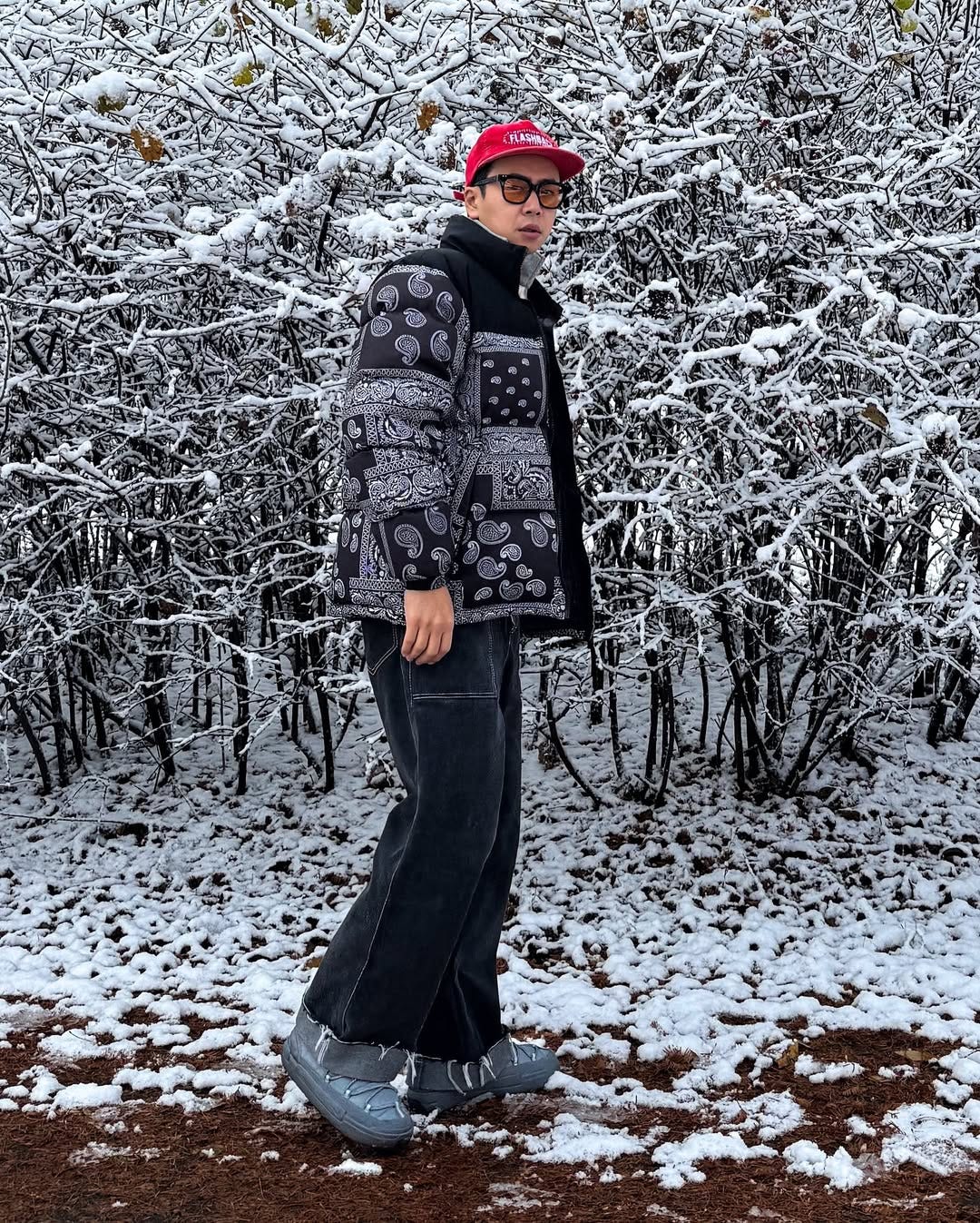 stylish man wearing a bandana print black and white puffer jacket with wide leg dark jeans and a red baseball hat. he's standing outside in front of trees that have icy snow on the branches