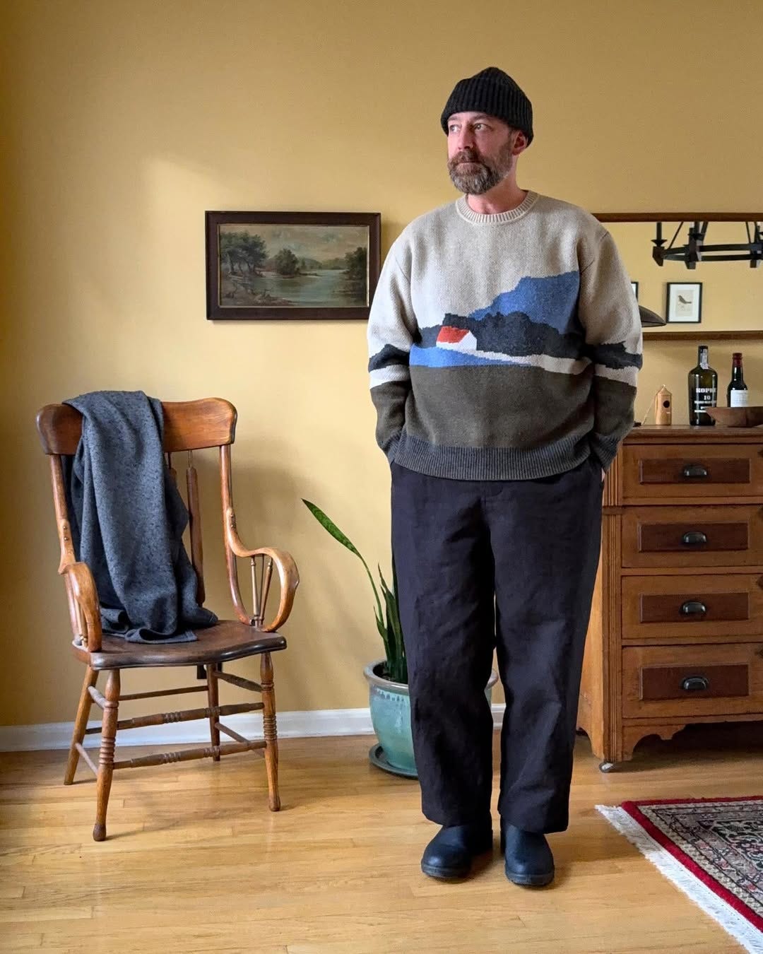 stylish man wearing a sweater with a landscape scene on the front with navy pants and black boots