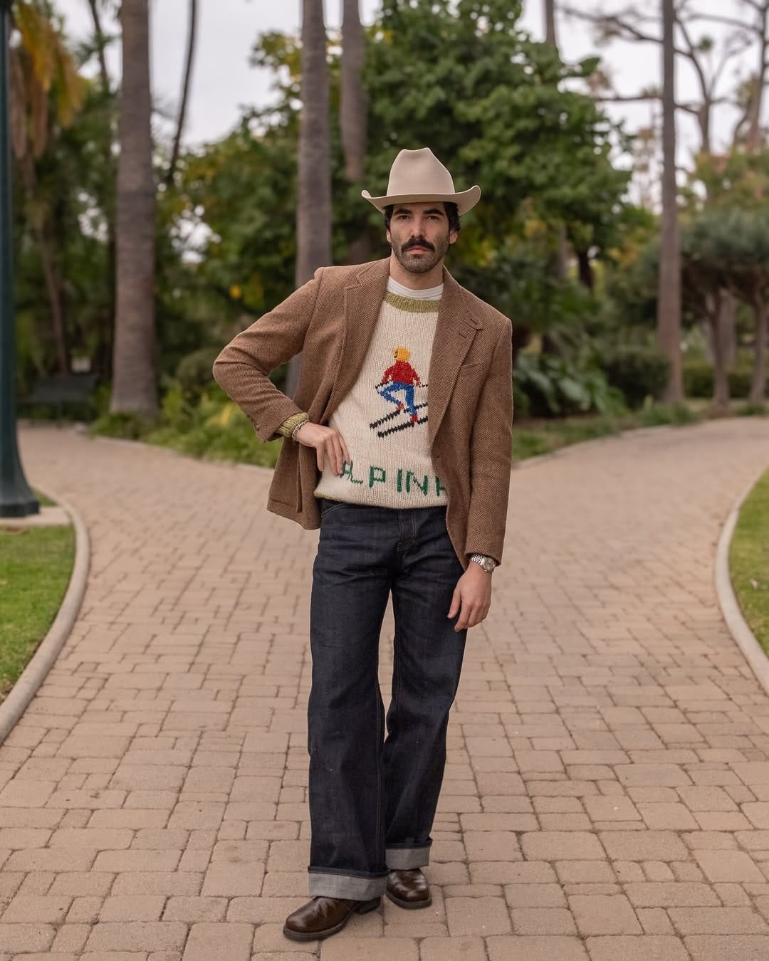 stylish man standing outside wearing a cowboy hat, brown jacket, sweater with an alpine skier on the front, dark jeans, and brown boots