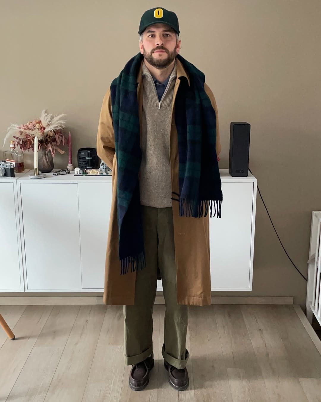 stylish man wearing a tan wool topcoat with a blackwatch scarf, over a quarter-zip sweater and chinos