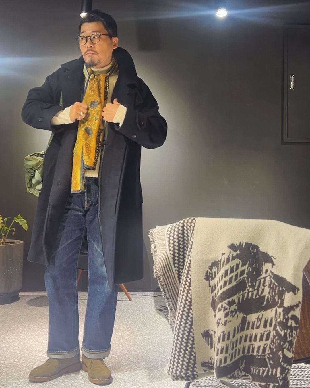 stylish man wearing a navy topcoat over a cream-colored turtleneck, dark jeans, boots, and a yellow silk scarf tied around his neck