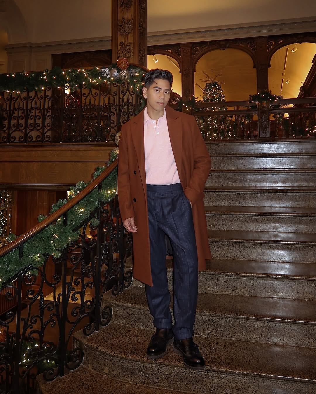 stylish young man standing on the steps of a hotel or restaurant decorated for the holidays, wearing a brown topcoat with a pink knit sweater and navy pinstripe pants